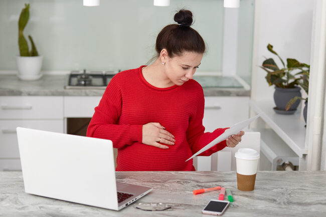 Attentive young pregnant female entrepreneur works from home, focused in document, wears casual clothes, studies data, works on laptop computer, drinks takeaway coffee, poses at kitchen table