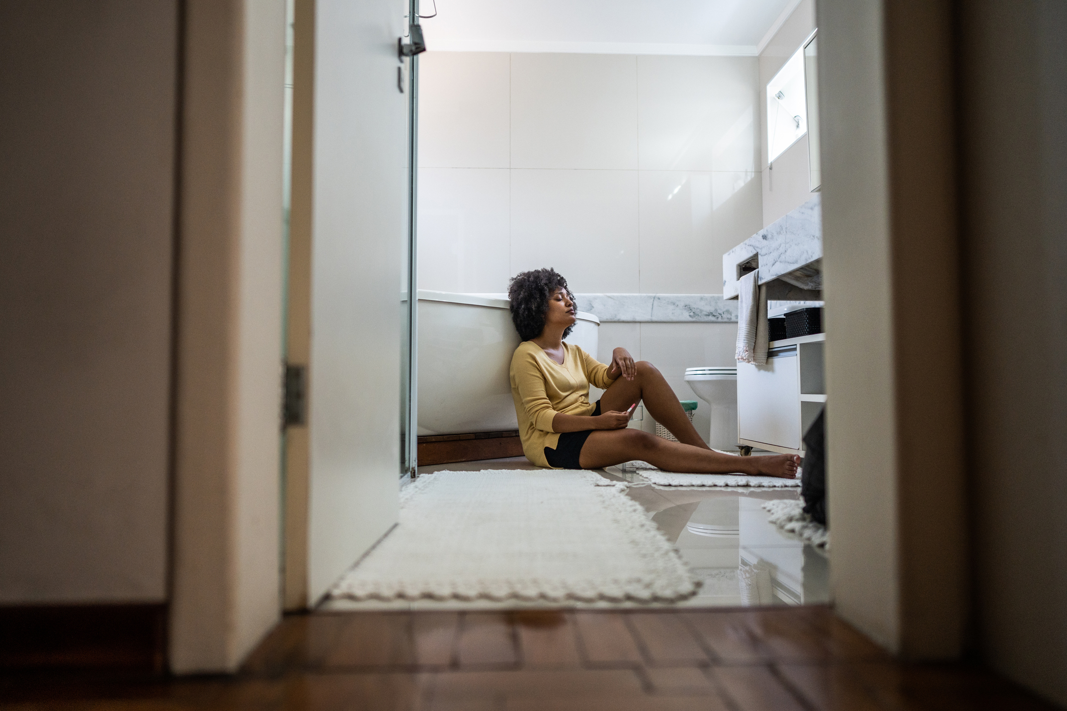 Sad young woman holding a pregnancy test sitting on ground in the bathroom at home